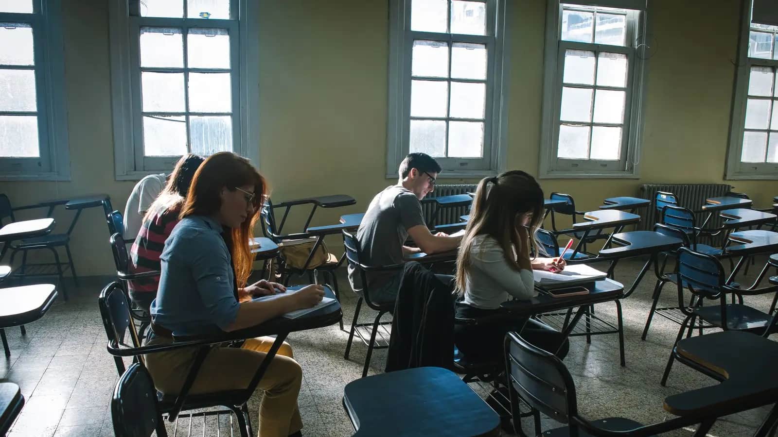 Students sitting at spaced desks writing their final school-leaving exams in a sunlit classroom