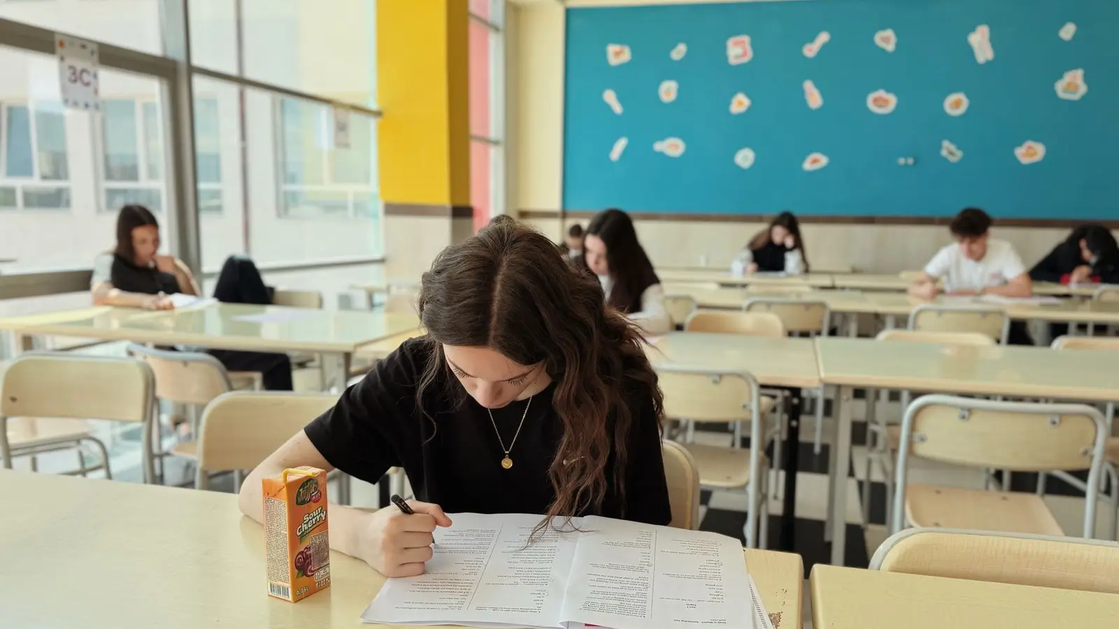 A teenage student concentrating on an exam paper at a classroom desk while classmates work at rows of desks behind